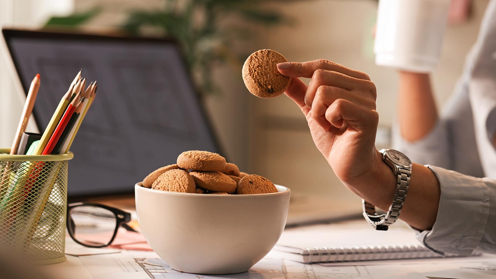 Eine Person sitzt an einem Schreibtisch, auf dem ein Laptop sowie eine Schüssel mit Cookies stehen.