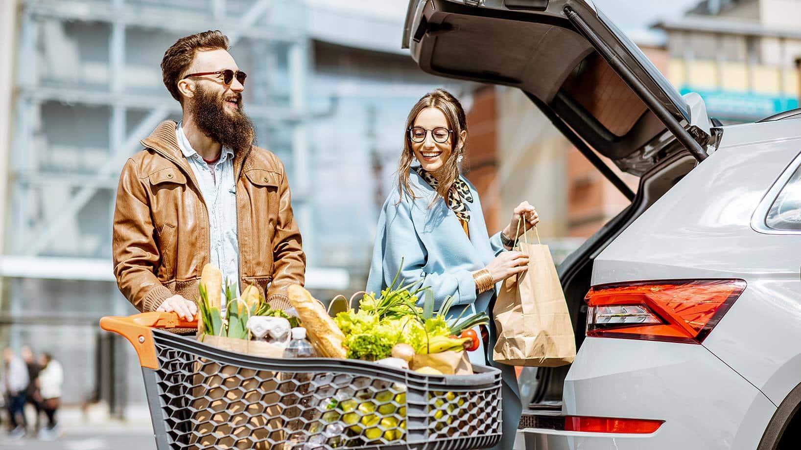 Auf dem Bild sind zwei Personen mit einem gefüllten Einkaufswagen zu sehen, die ihre Einkäufe in ein Auto laden.